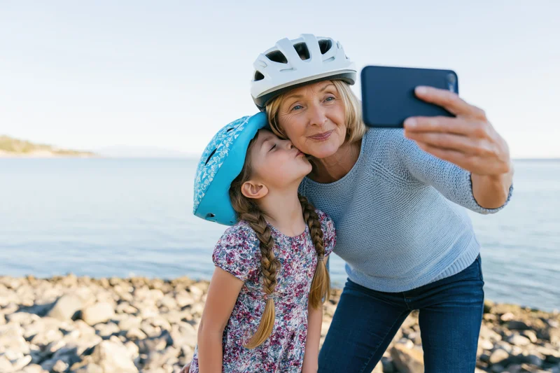 Grandmother and granddaughter taking a selfie by the water, both wearing helmets, the girl kissing her cheek