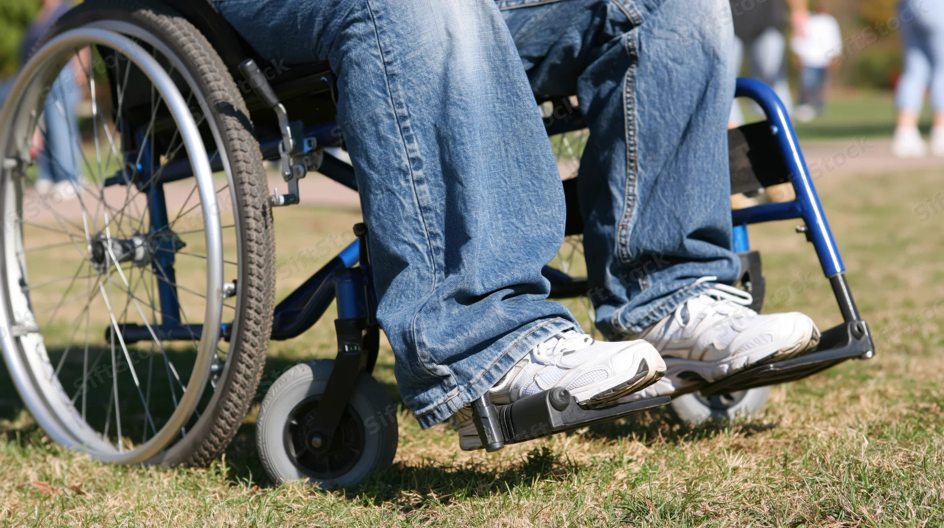 Person in wheelchair with legs in jeans and sneakers resting on footrest in grassy park area