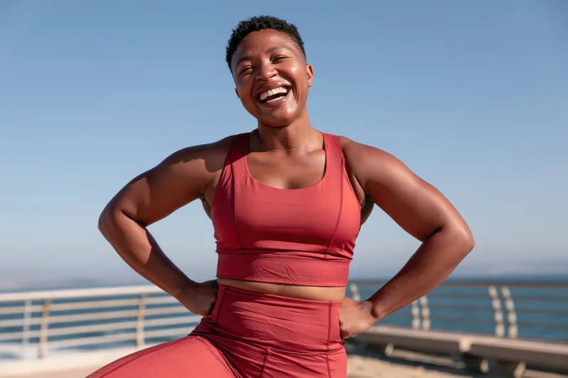 Smiling black woman in red sportswear posing with hands on hips by ocean railing