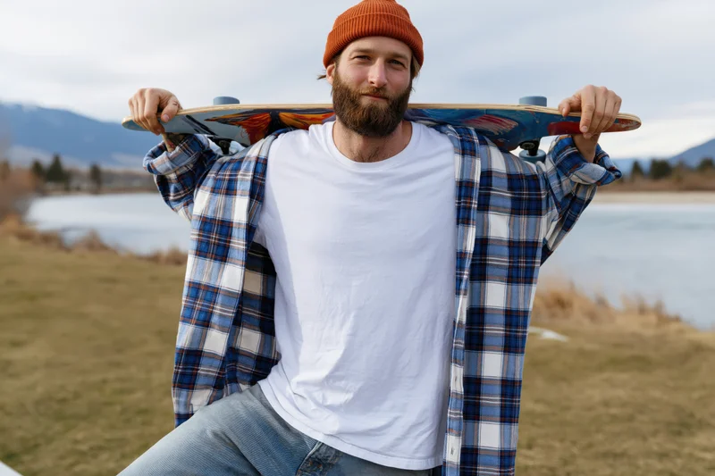 Bearded man holding skateboard over shoulder outdoors near lake and mountains
