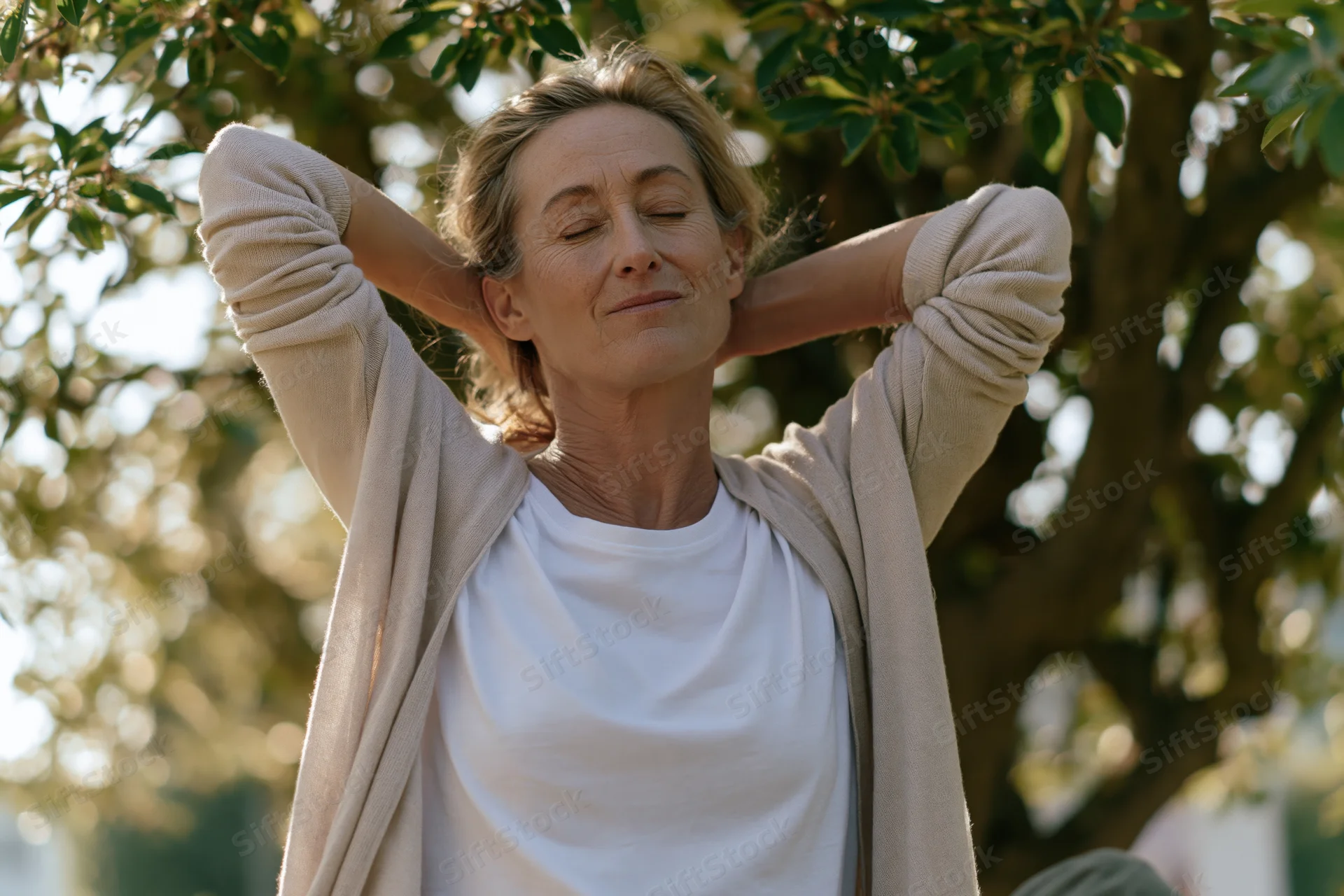 Middle-aged woman stretching neck with hands behind head outdoors in garden