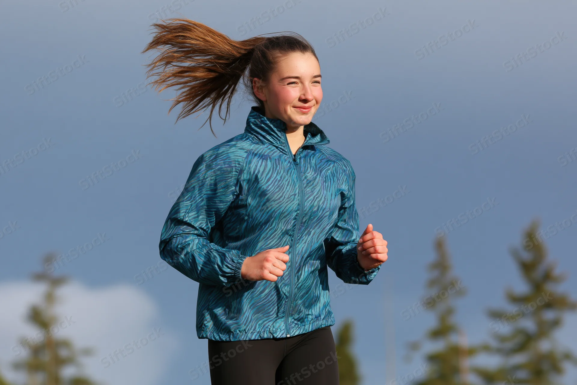 Young woman running outdoors in a teal jacket with hair flying in the wind among pine trees