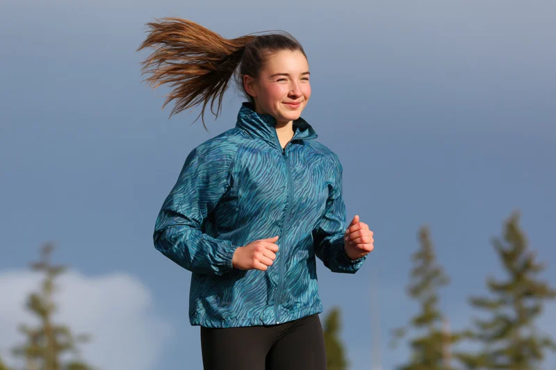 Young woman running outdoors in a teal jacket with hair flying in the wind among pine trees