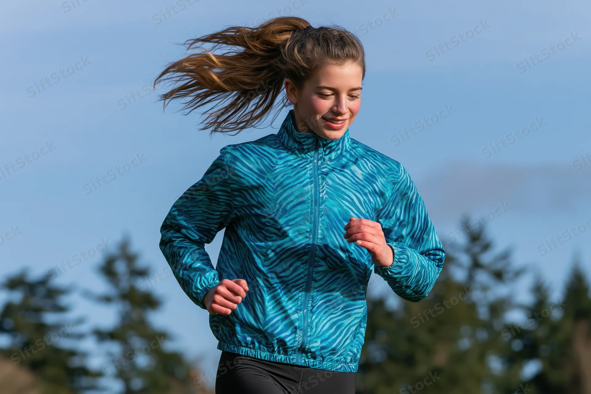 Young woman running outdoors in a teal patterned jacket with hair in motion against trees and sky