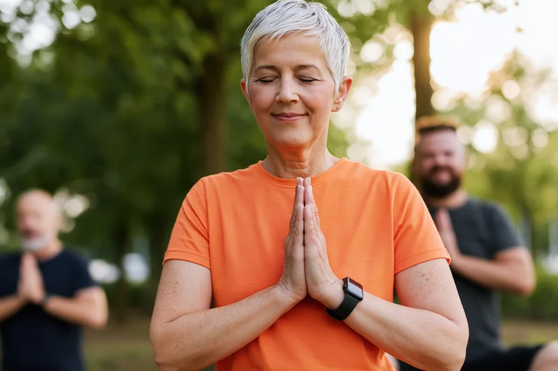 Woman in prayer position meditating outdoors in a park with others in the background