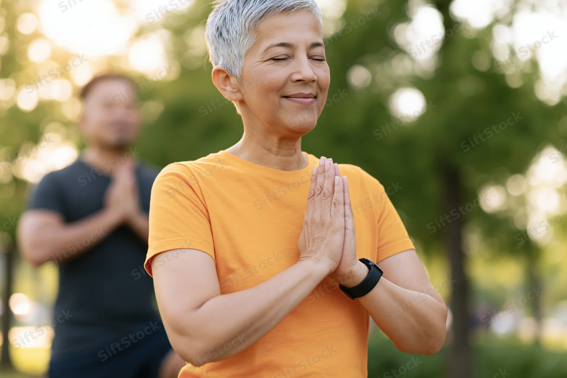 Woman practicing yoga with hands together in a park, eyes closed and smiling, with a man in the background also meditating