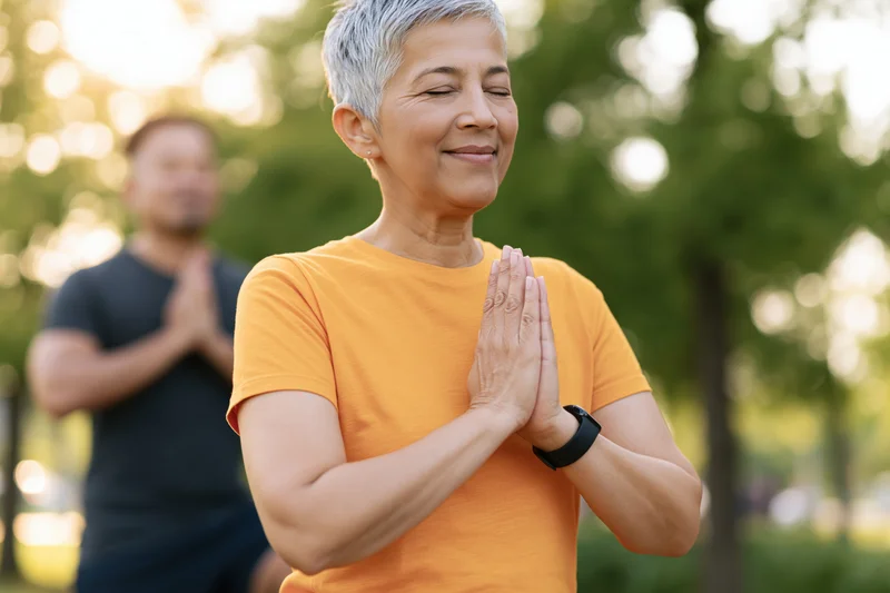 Woman practicing yoga with hands together in a park, eyes closed and smiling, with a man in the background also meditating