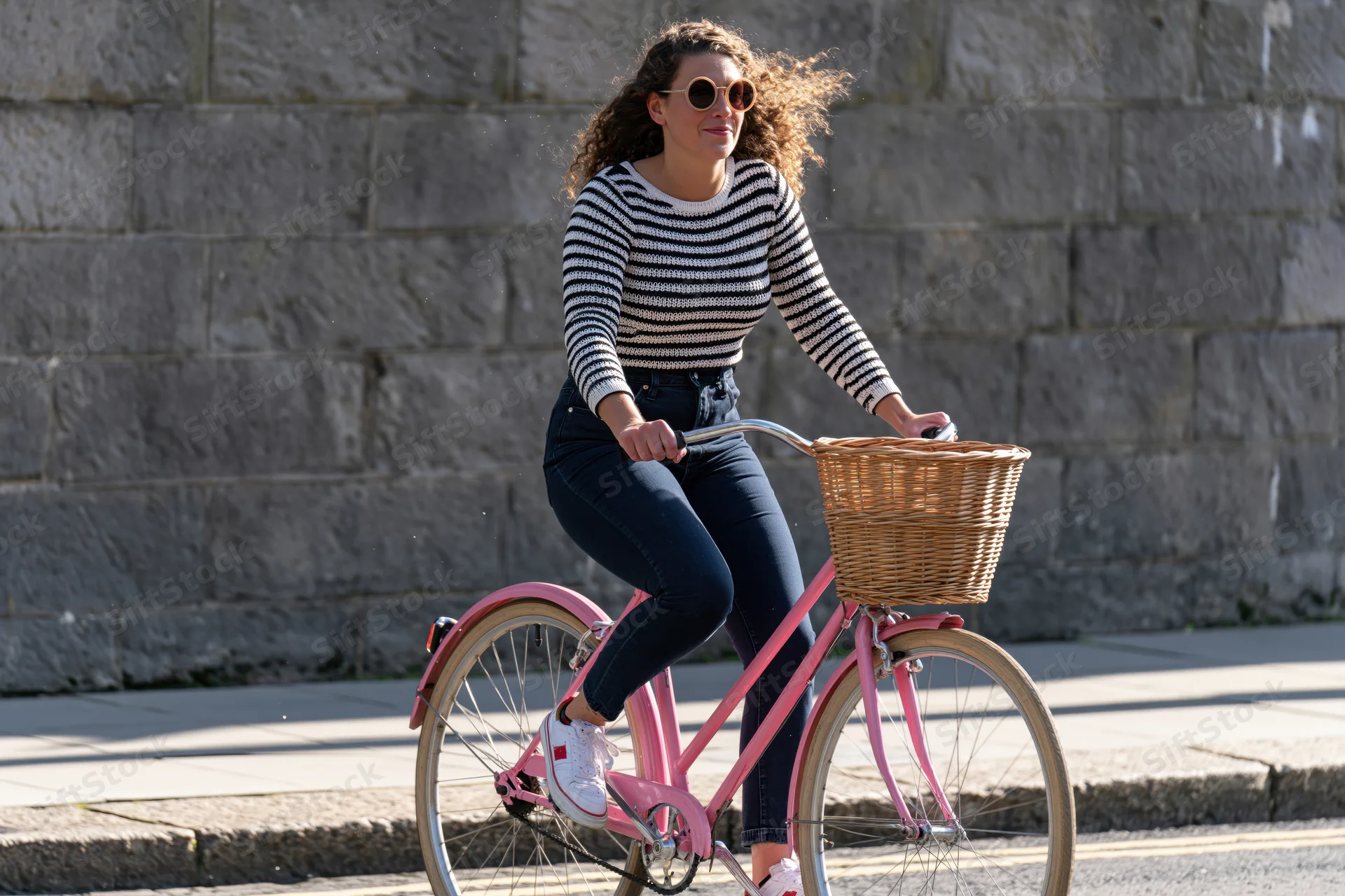 Woman riding a pink bicycle with a wicker basket along a city sidewalk