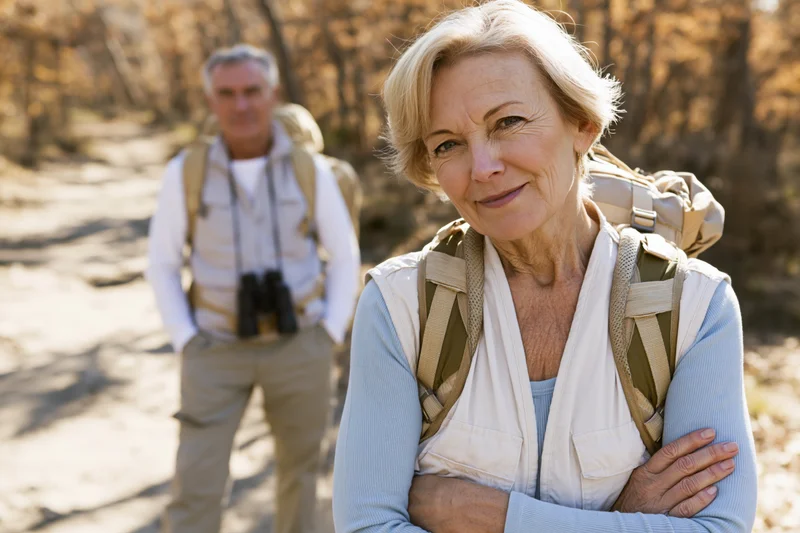 Senior woman smiling with arms crossed while hiking on a forest trail, man with binoculars behind her
