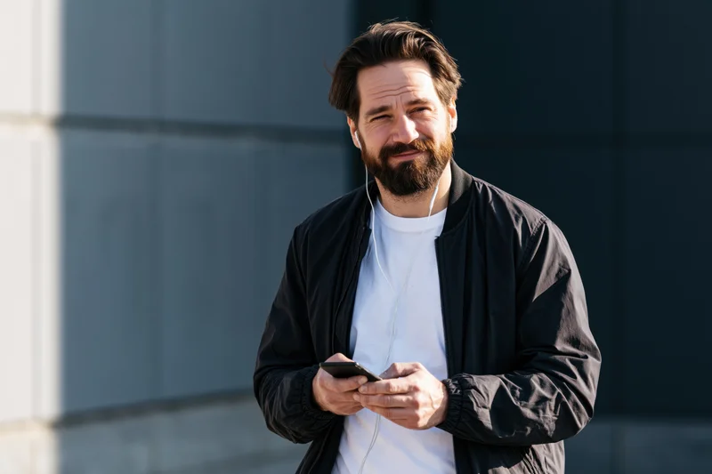Bearded man smiling while using smartphone outdoors in modern urban setting