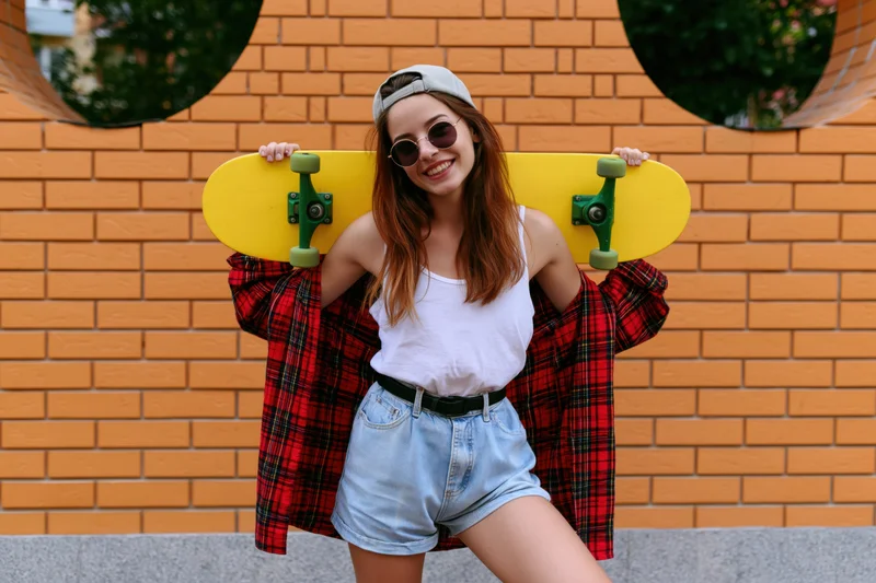 Young woman smiling while holding a yellow skateboard against an orange brick wall