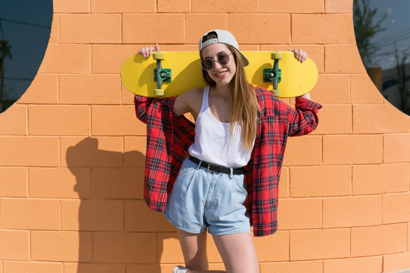 Young woman holding yellow skateboard against orange brick wall with circular cutouts, smiling in casual streetwear outfit