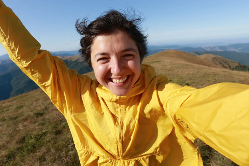 Woman smiling in yellow jacket on mountain summit with arms raised, overlooking valley and hills
