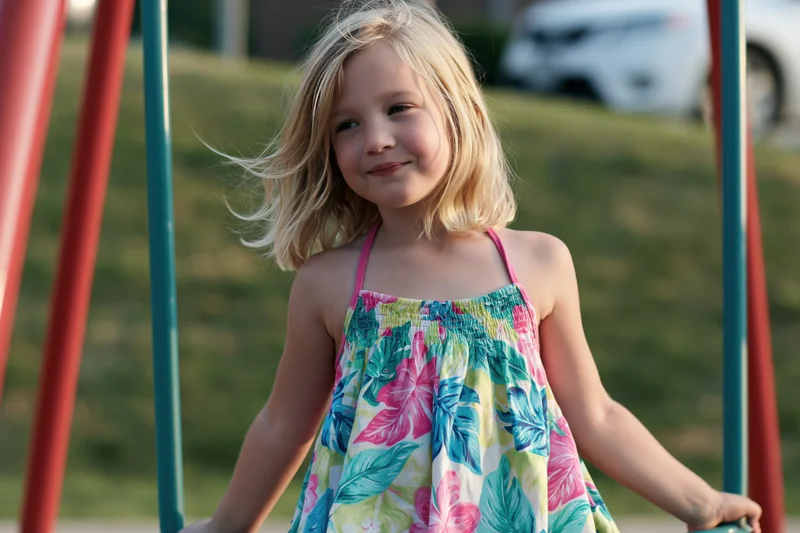 Young girl smiling on a playground swing in a floral dress during summer day