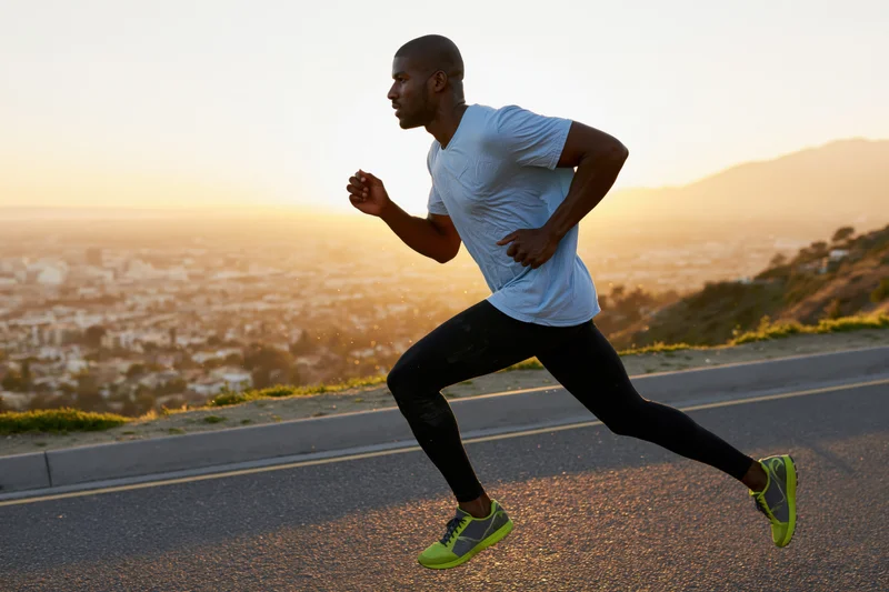 Fit black man running mid-stride on paved road overlooking city at sunrise