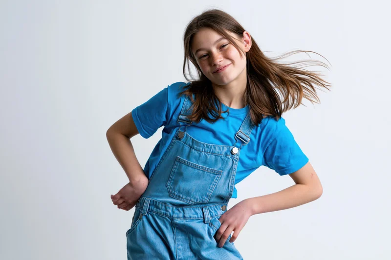 Girl in blue denim overalls and tshirt posing mid-tilt with hands on hips against white background