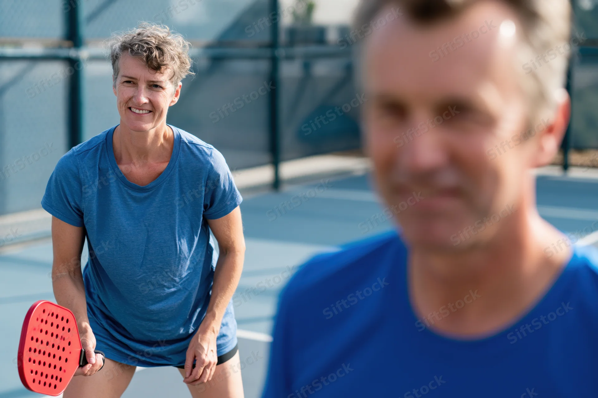 Woman holding red paddle smiling while playing pickleball on outdoor court with man in foreground