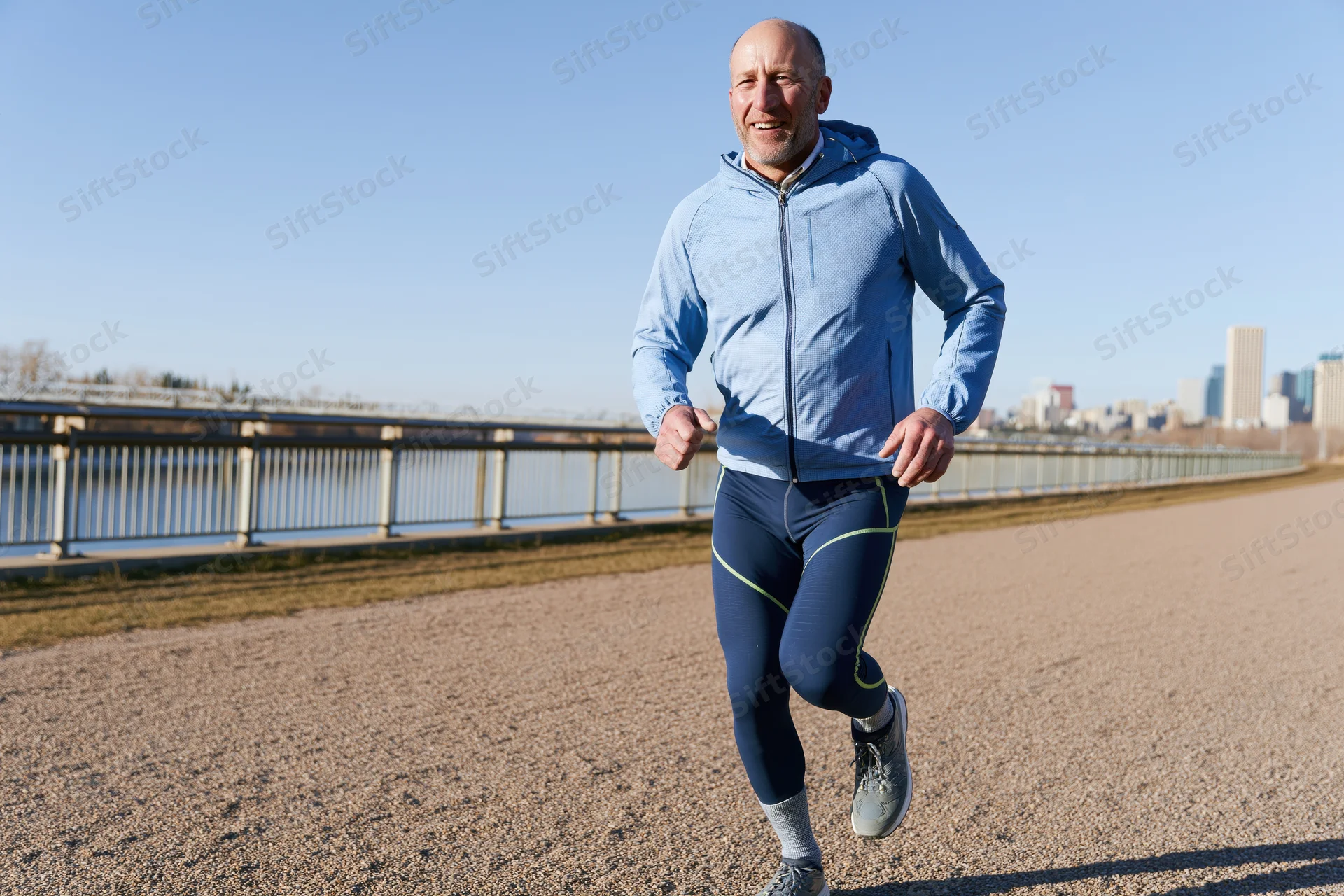 Bald man running on gravel path by river with city skyline in background