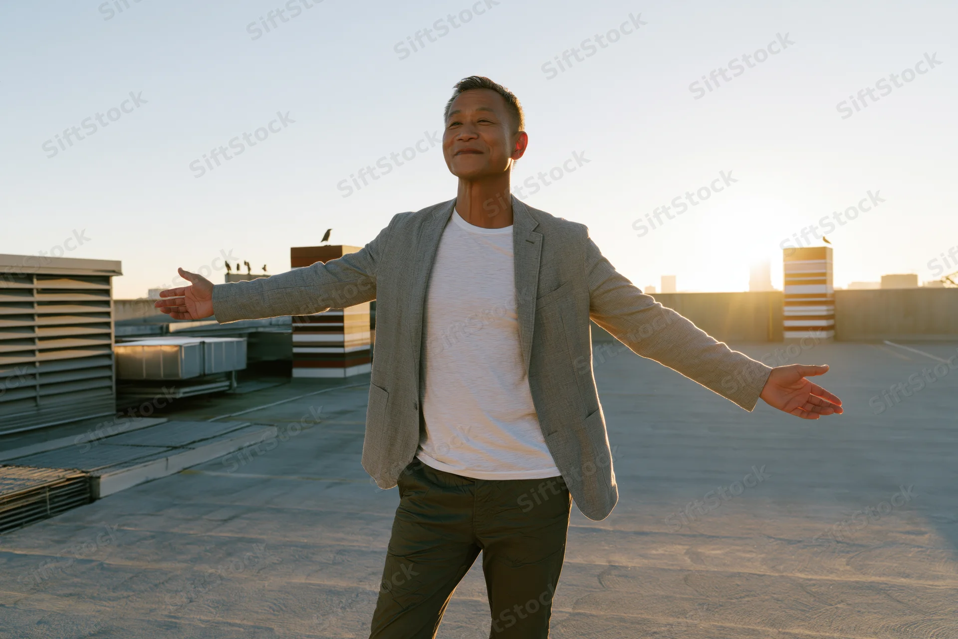 Middle-aged asian man standing on rooftop with arms open facing sunset city skyline