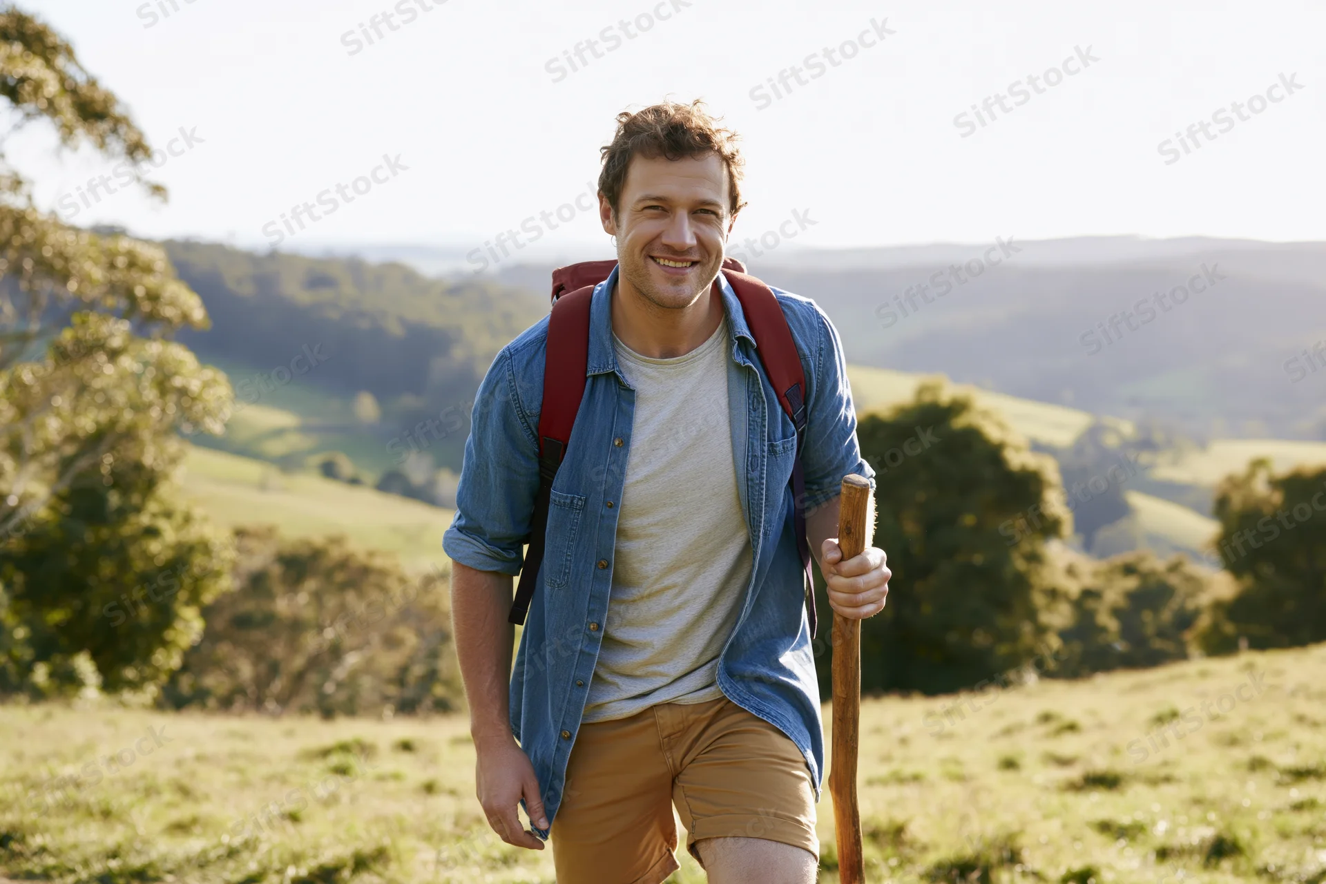 Smiling man hiking with backpack and wooden stick in sunlit countryside