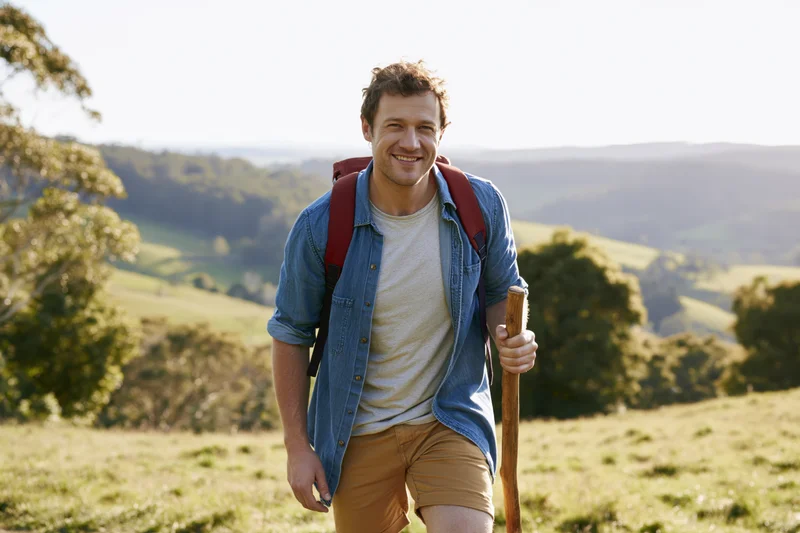 Smiling man hiking with backpack and wooden stick in sunlit countryside