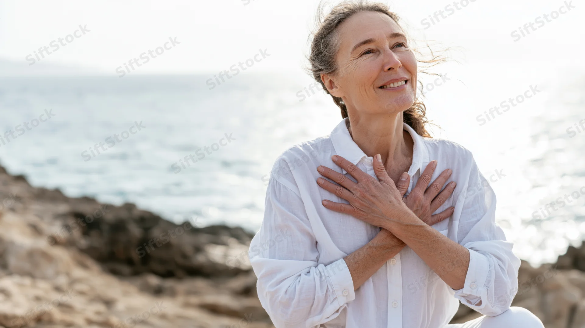 Senior woman smiling with hands on chest by the ocean on a windy day