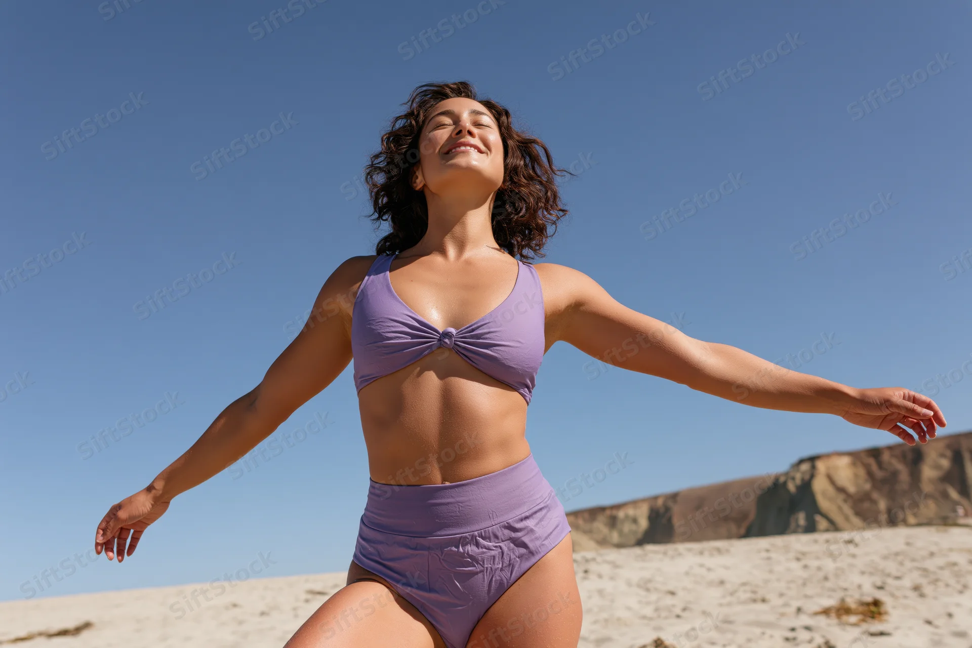 Woman in purple bikini smiling with arms outstretched on sunny beach
