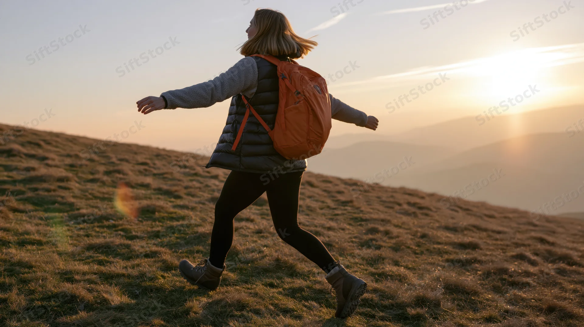 Woman hiking on a grassy hill at sunset with backpack, arms outstretched
