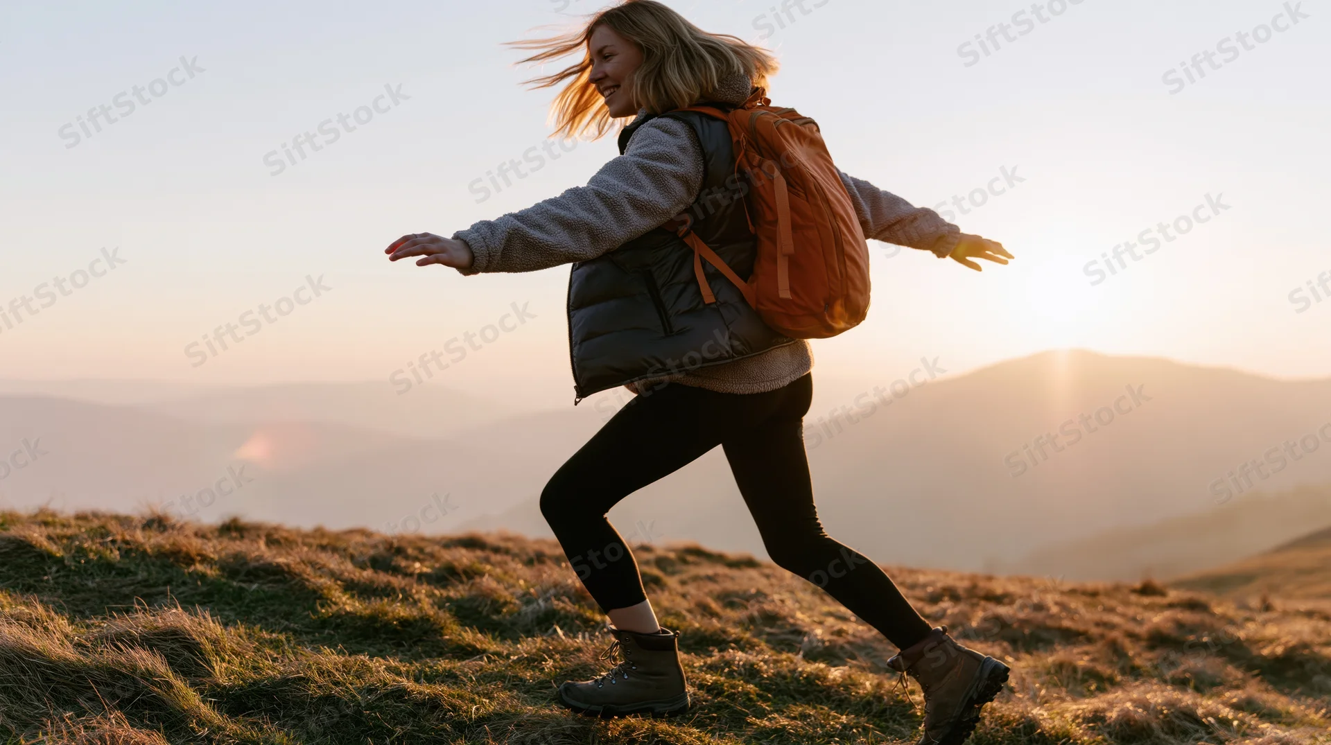 Woman hiking on a grassy mountain trail at sunset, arms outstretched, smiling, wearing backpack and outdoor gear
