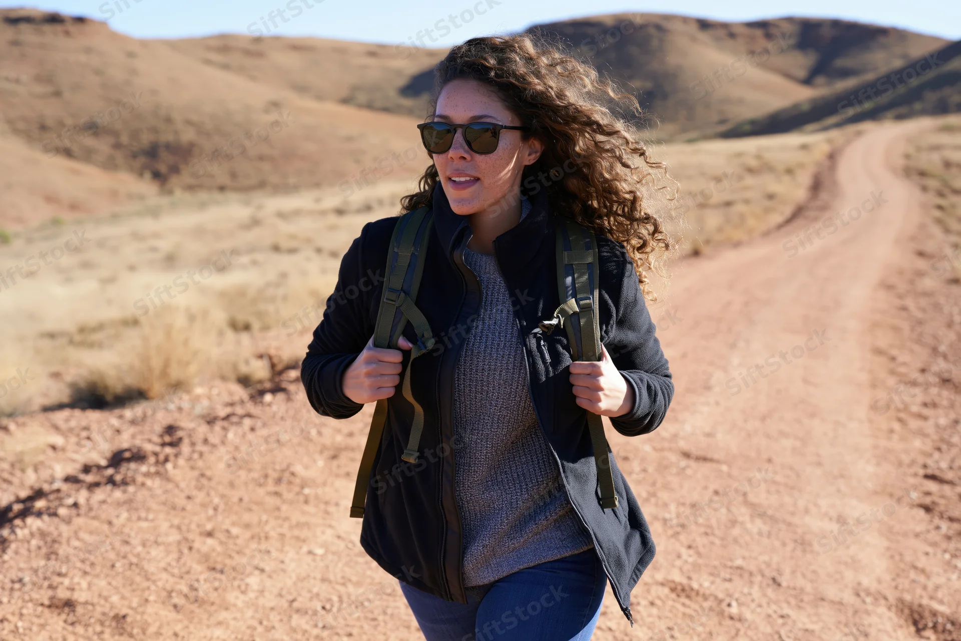 Woman hiking on a dirt trail in a desert landscape wearing sunglasses and a backpack