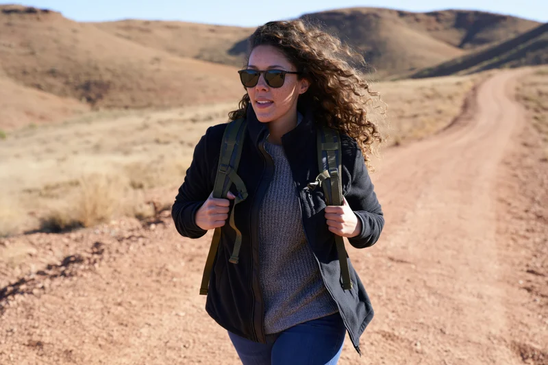 Woman hiking on a dirt trail in a desert landscape wearing sunglasses and a backpack