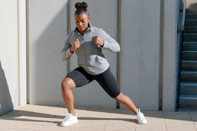Young black woman performing squats in sportswear on urban pavement near stairs