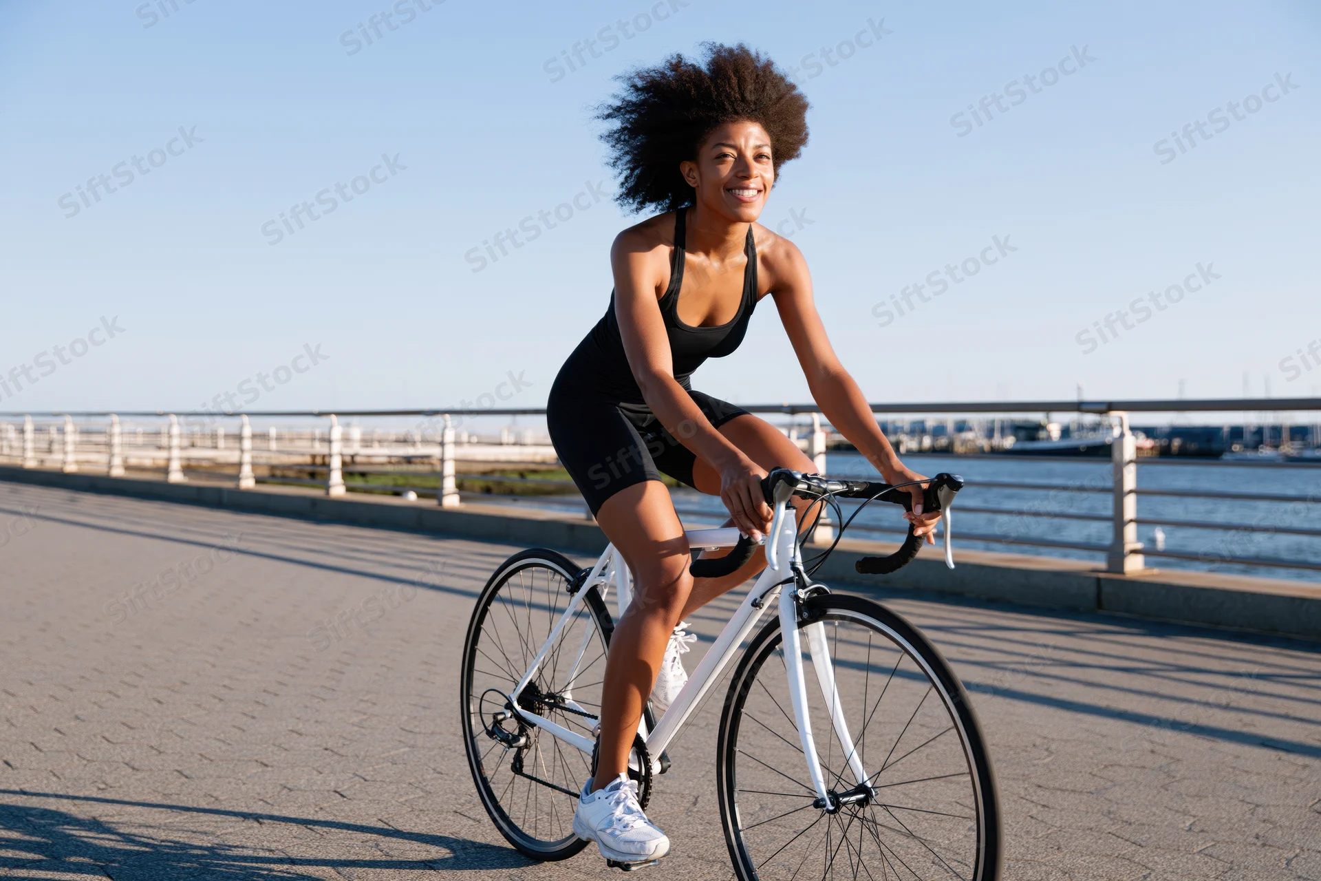 Young black woman cycling on paved path by waterfront smiling in motion