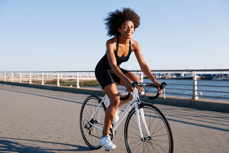 Young black woman cycling on paved path by waterfront smiling in motion