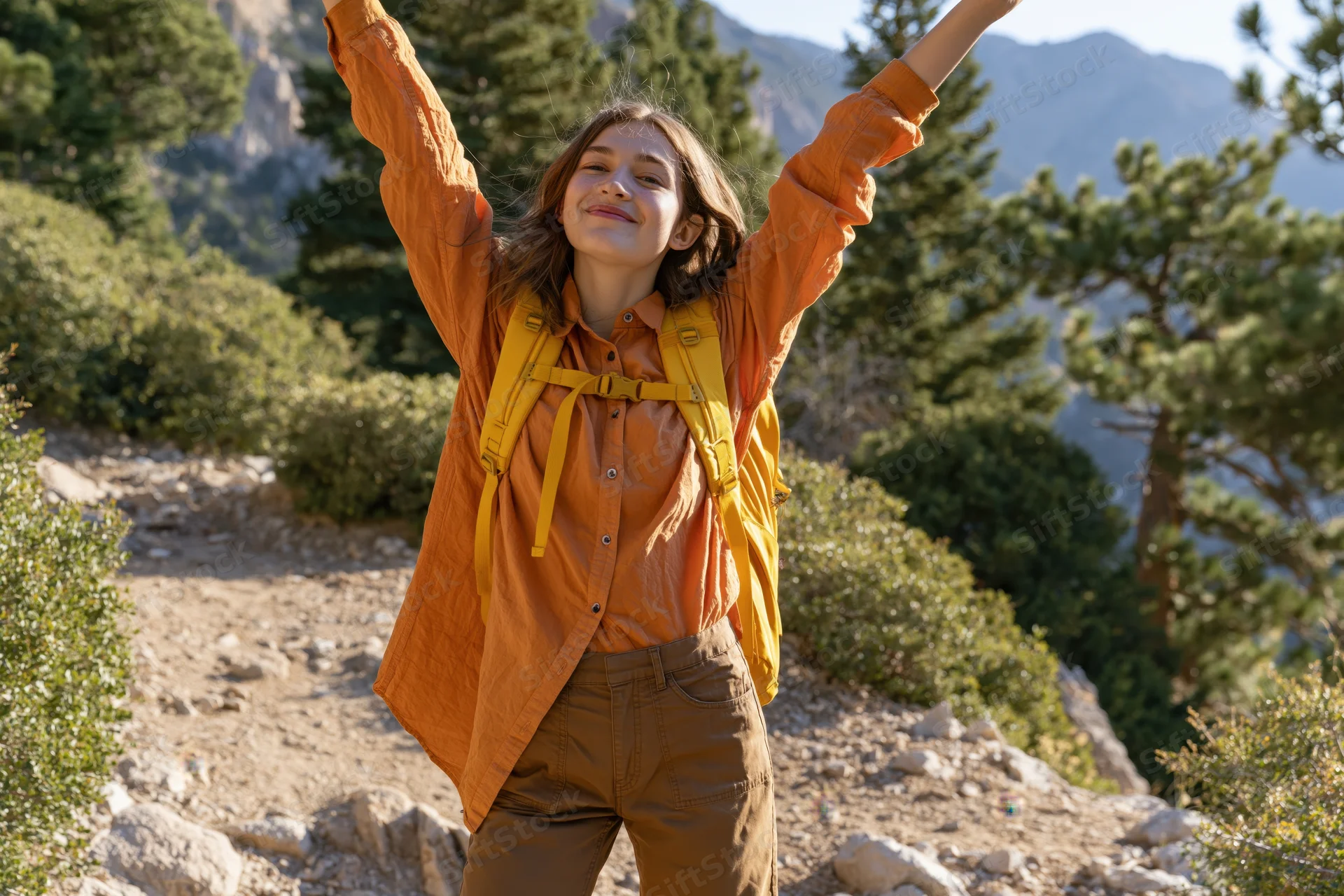 Young woman with arms raised smiling on a mountain trail wearing a backpack and orange shirt