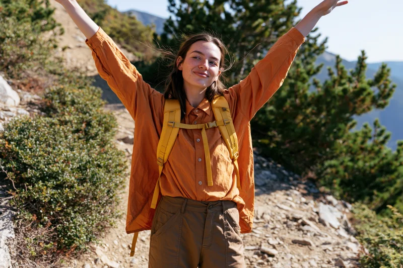 Young woman with arms raised smiling on a mountain trail wearing a backpack