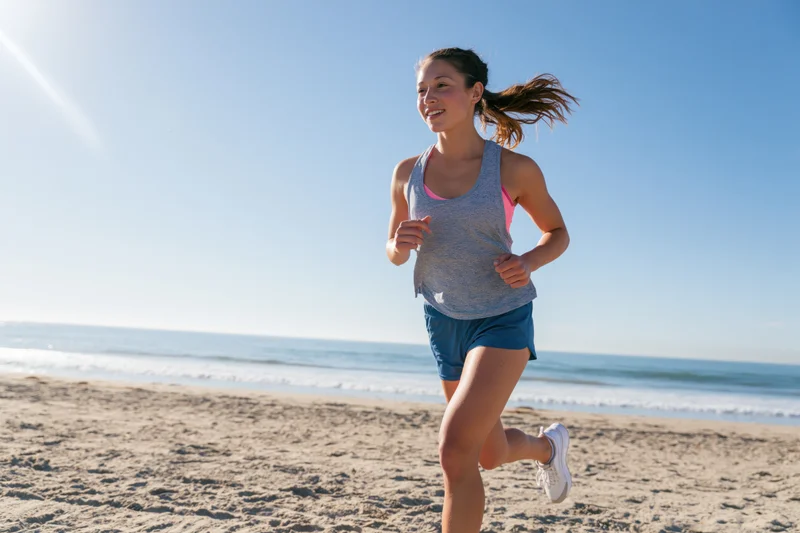 Young woman running on sandy beach near ocean under clear blue sky