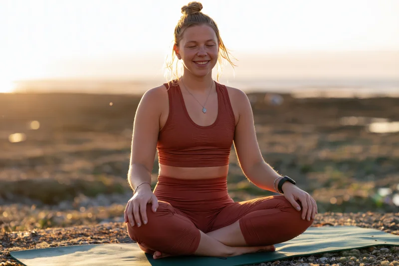 Young woman meditating cross-legged on a yoga mat at sunset on a gravel beach