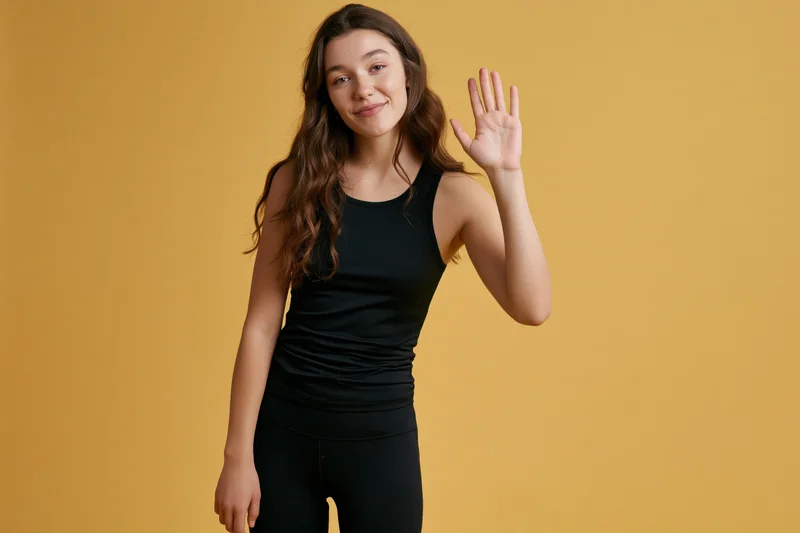 Young woman waving and smiling in black activewear against yellow background