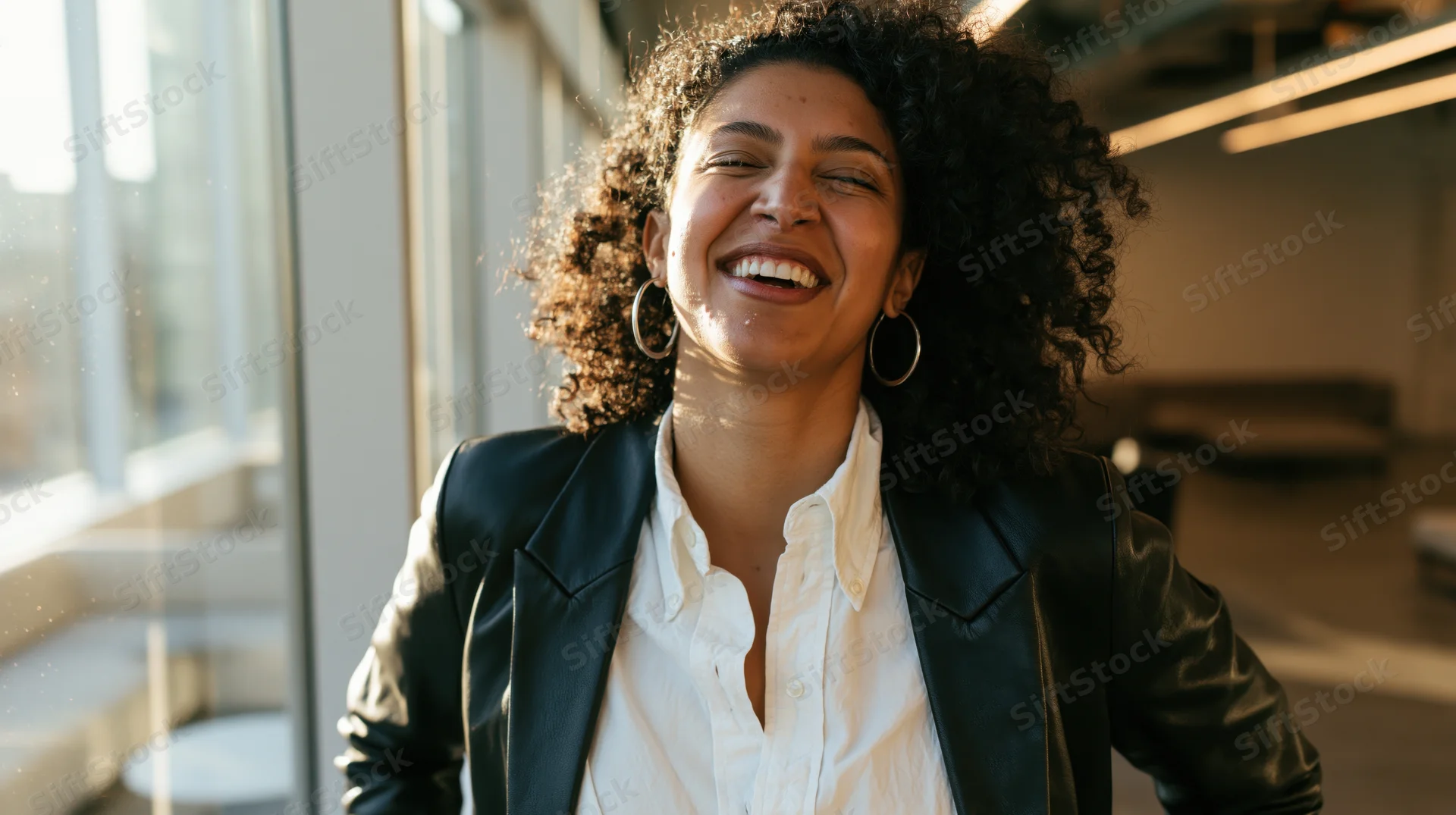 Young woman laughing joyfully in modern office near window with sunlight streaming in
