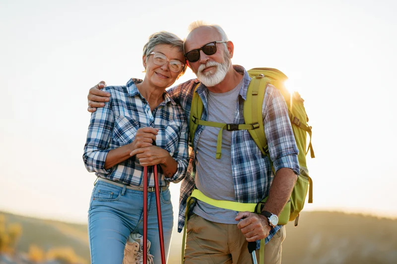 Senior couple hiking together outdoors with backpack and trekking poles at sunset