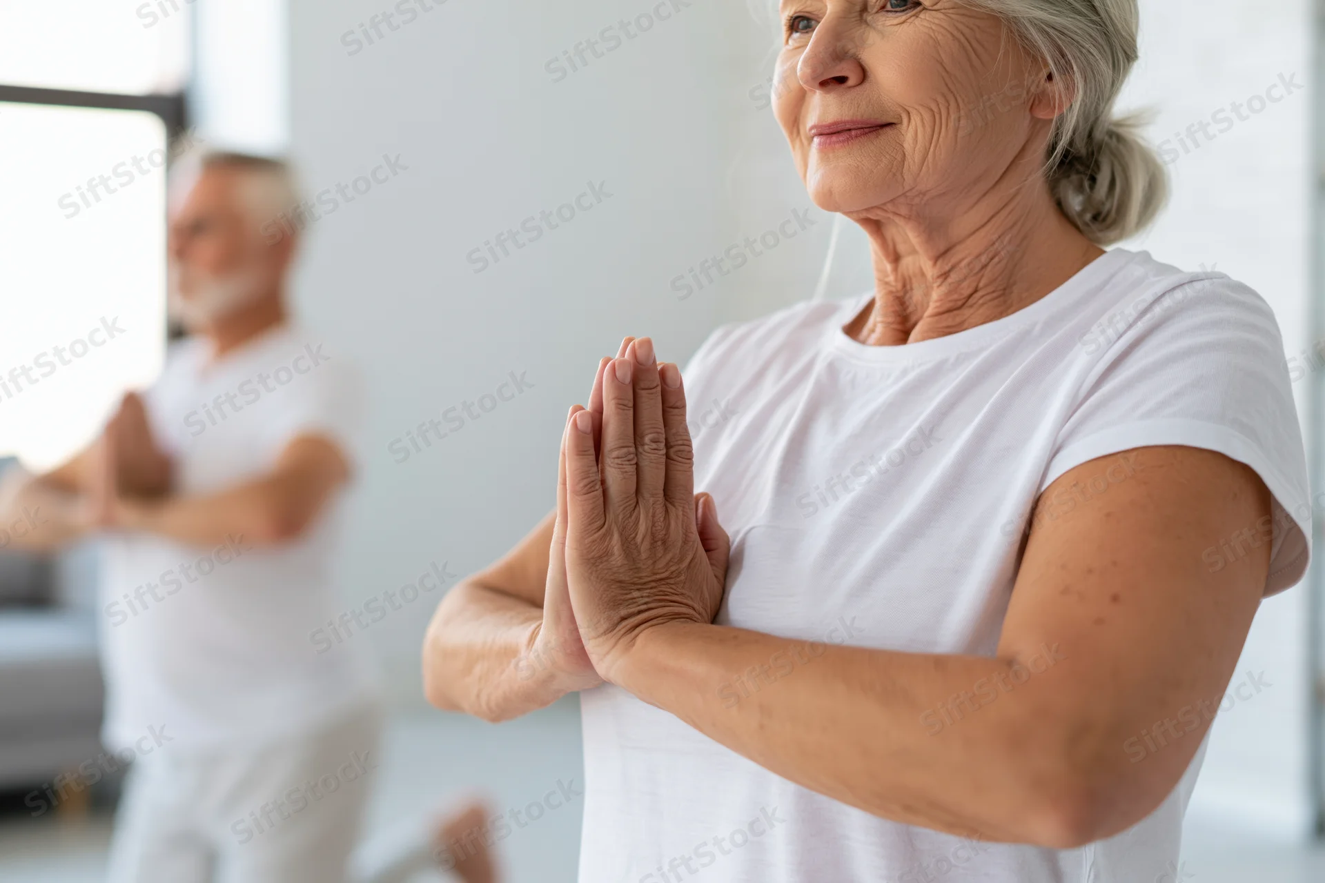 Senior woman in white tshirt holding prayer pose during yoga practice with older man in background