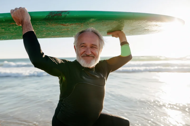 Senior man smiling while carrying surfboard on beach at sunrise