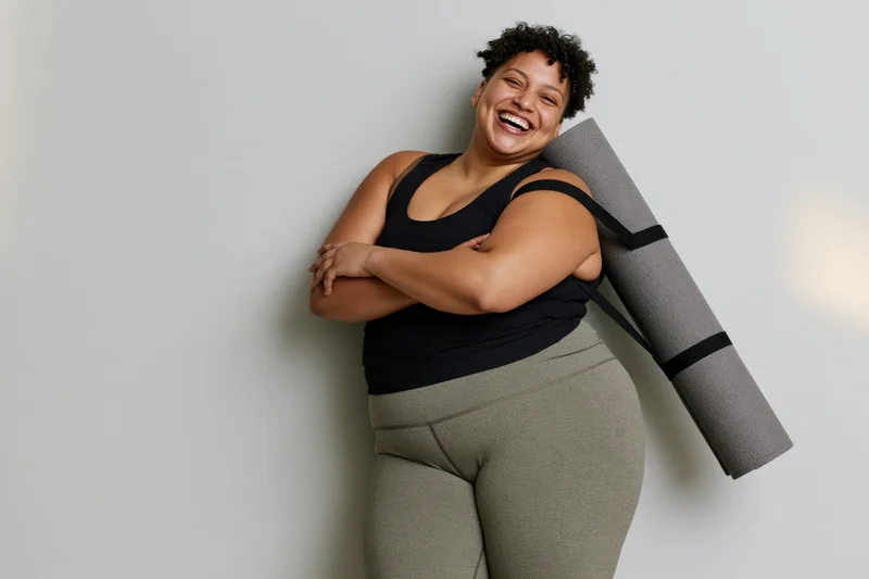 Plus-size woman laughing with yoga mat over shoulder leaning against wall