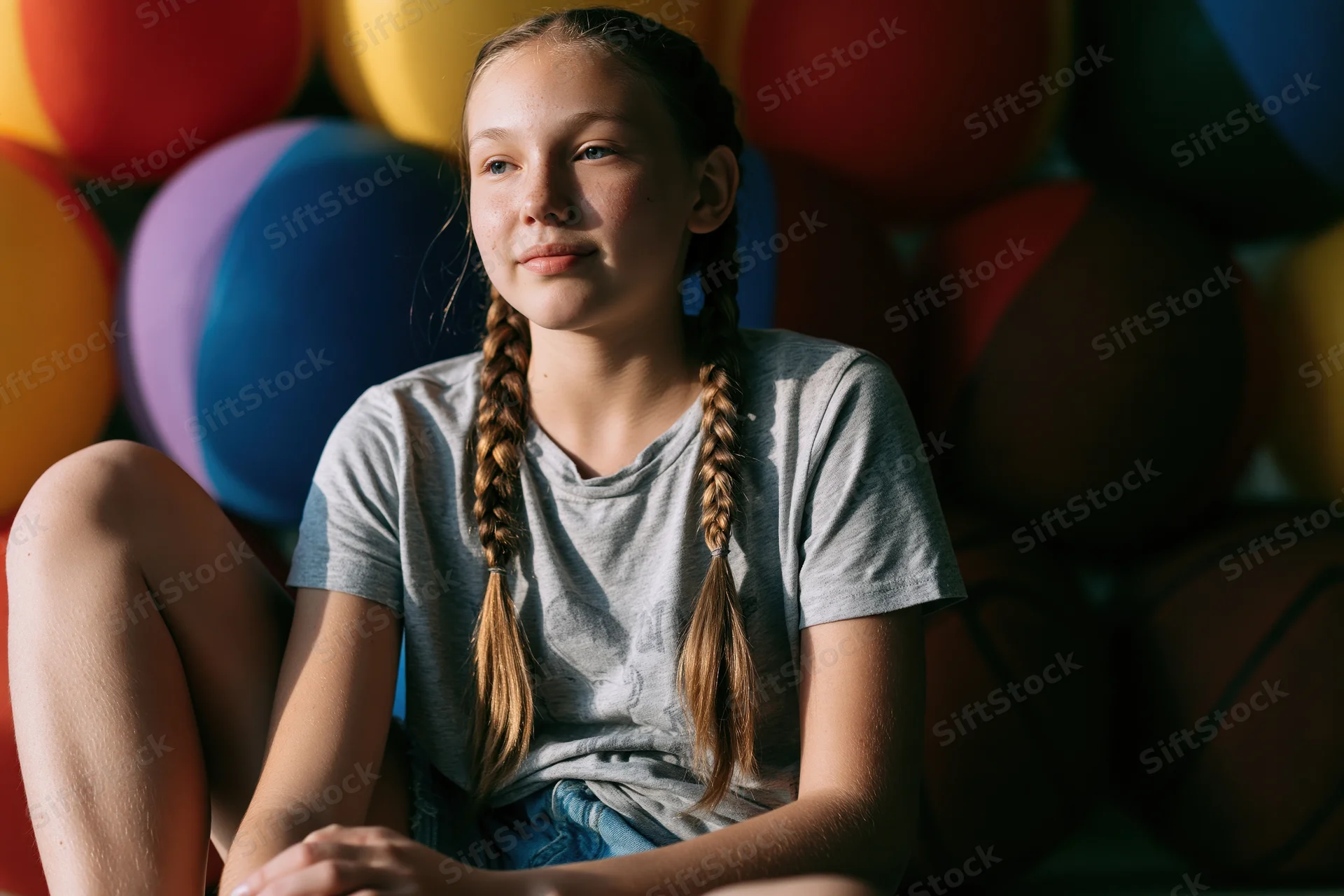 Young girl sitting with braids and casual clothes surrounded by colorful balloons indoors