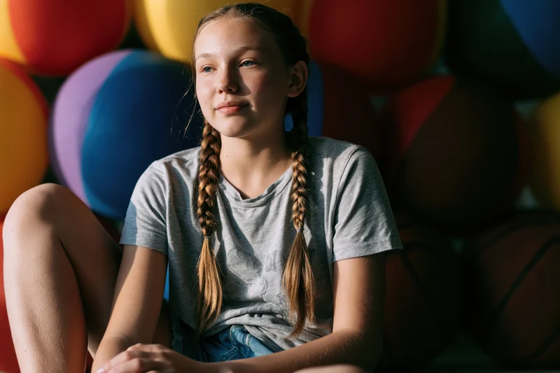 Young girl sitting with braids and casual clothes surrounded by colorful balloons indoors