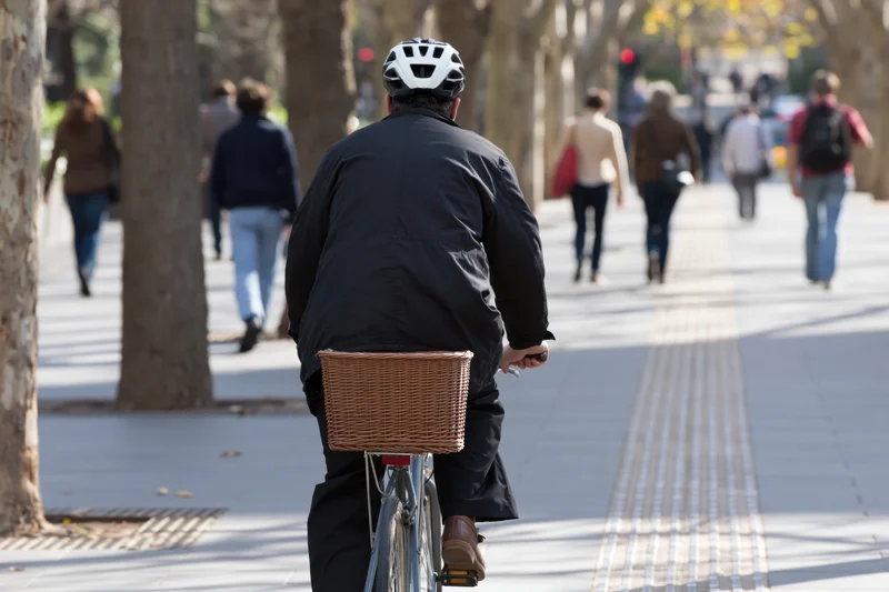 Cyclist riding bicycle with wicker basket on city sidewalk lined with trees, seen from behind with pedestrians walking ahead
