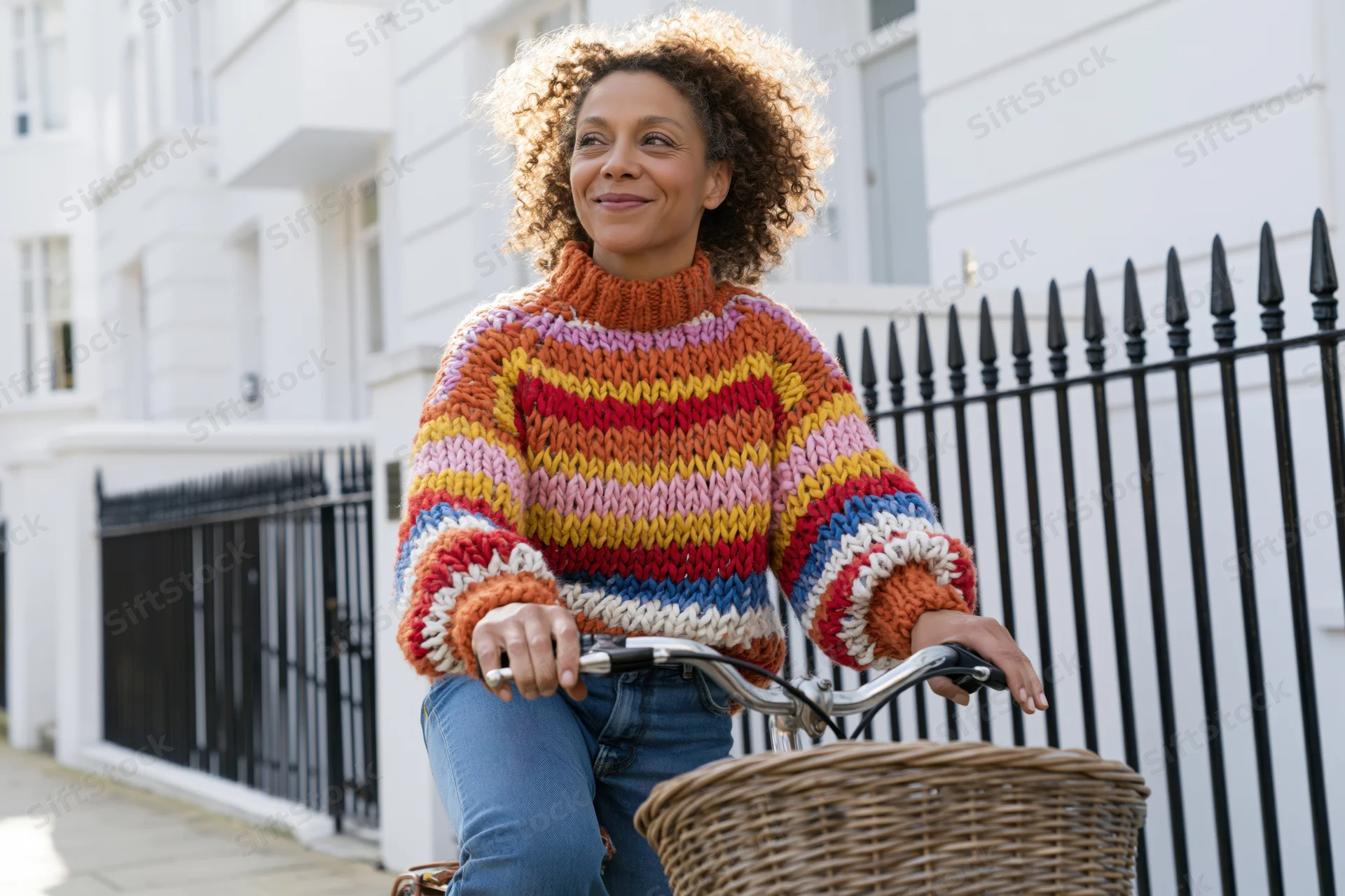 Woman riding bicycle on urban sidewalk with colorful knitted sweater and wicker basket