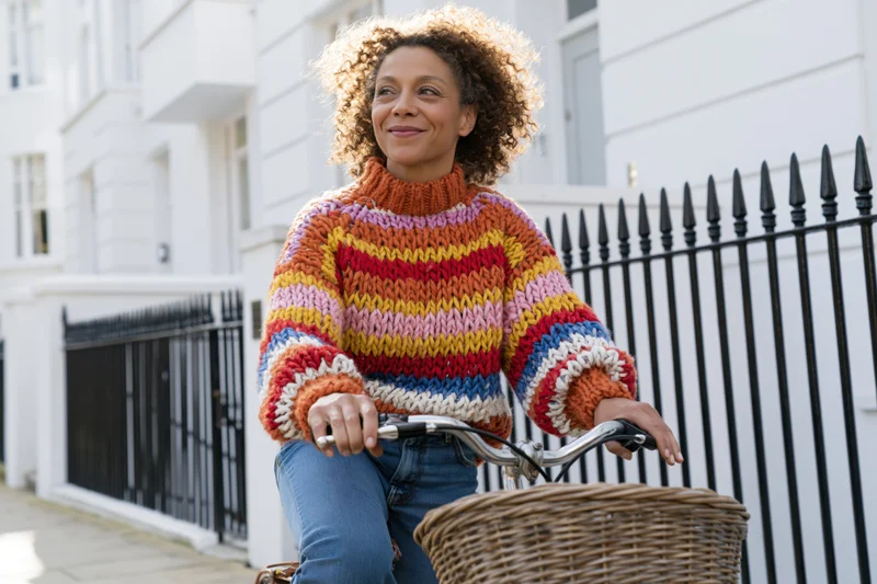 Woman riding bicycle on urban sidewalk with colorful knitted sweater and wicker basket