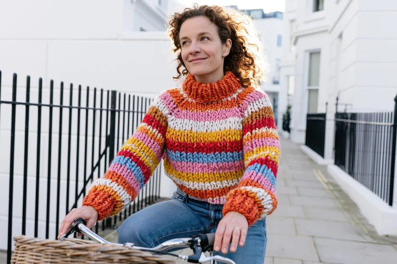 Woman riding bicycle on urban sidewalk wearing colorful knitted sweater and jeans